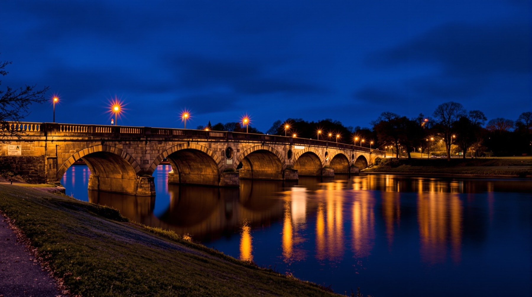 A stone river bridge lit at dusk with reflections in the water below