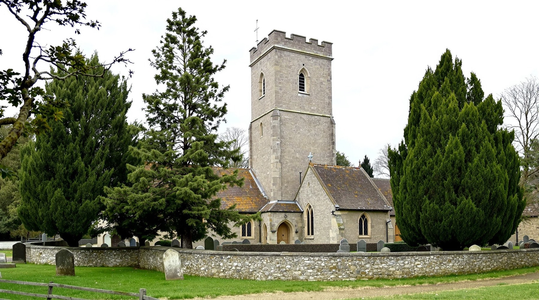 An old English village church with a tall square stone tower behind a graveyard wall